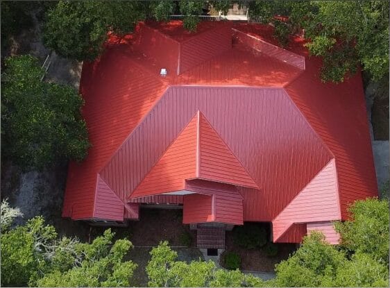 Aerial view of house with distinctive red metal roof surrounded by green trees